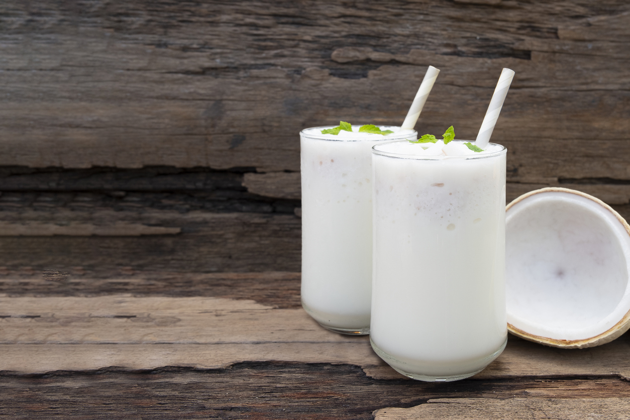Zwei hohe Gläser Kokos-Milchshake mit Strohhalmen und Minzblättern vor halber Kokosnuss.