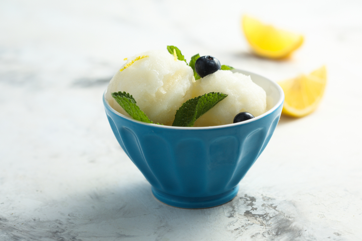 Zwei Kugeln Zitronensorbet mit Minze und Blaubeeren in blauer Schale, hinten Zitronenspalten.