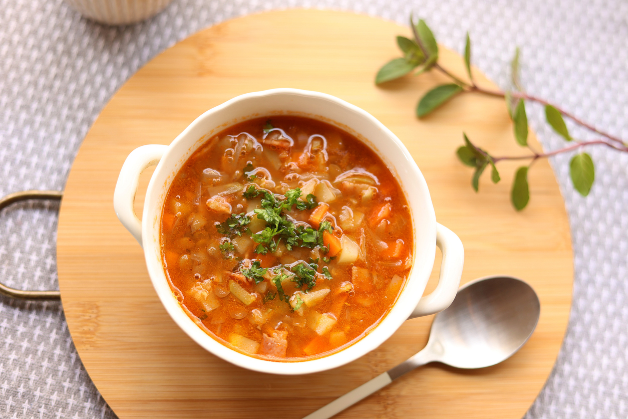 Draufsicht auf eine weiße Schale mit tomatiger Linsen-Minestrone und grüner Petersilie, daneben ein Löffel.