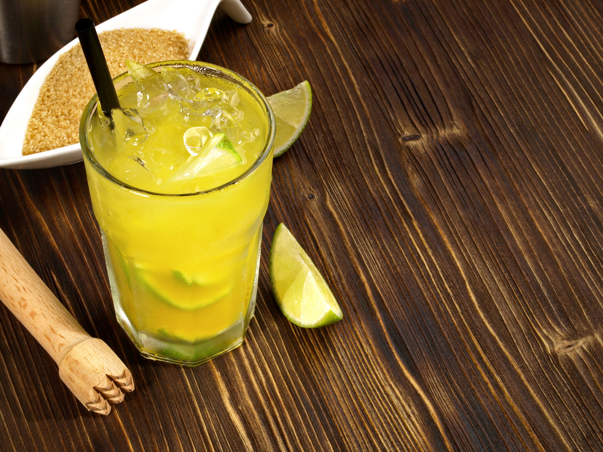 Gelber Ipanema-Mocktail im Glas mit Eiswürfeln, Limettenspalten und Strohhalm auf Holztisch.