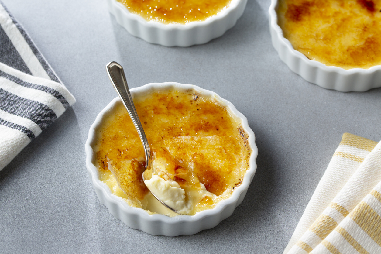 Weiße Crème-brûlée im Ramekin, ein Löffel bricht die karamellisierte Oberfläche.