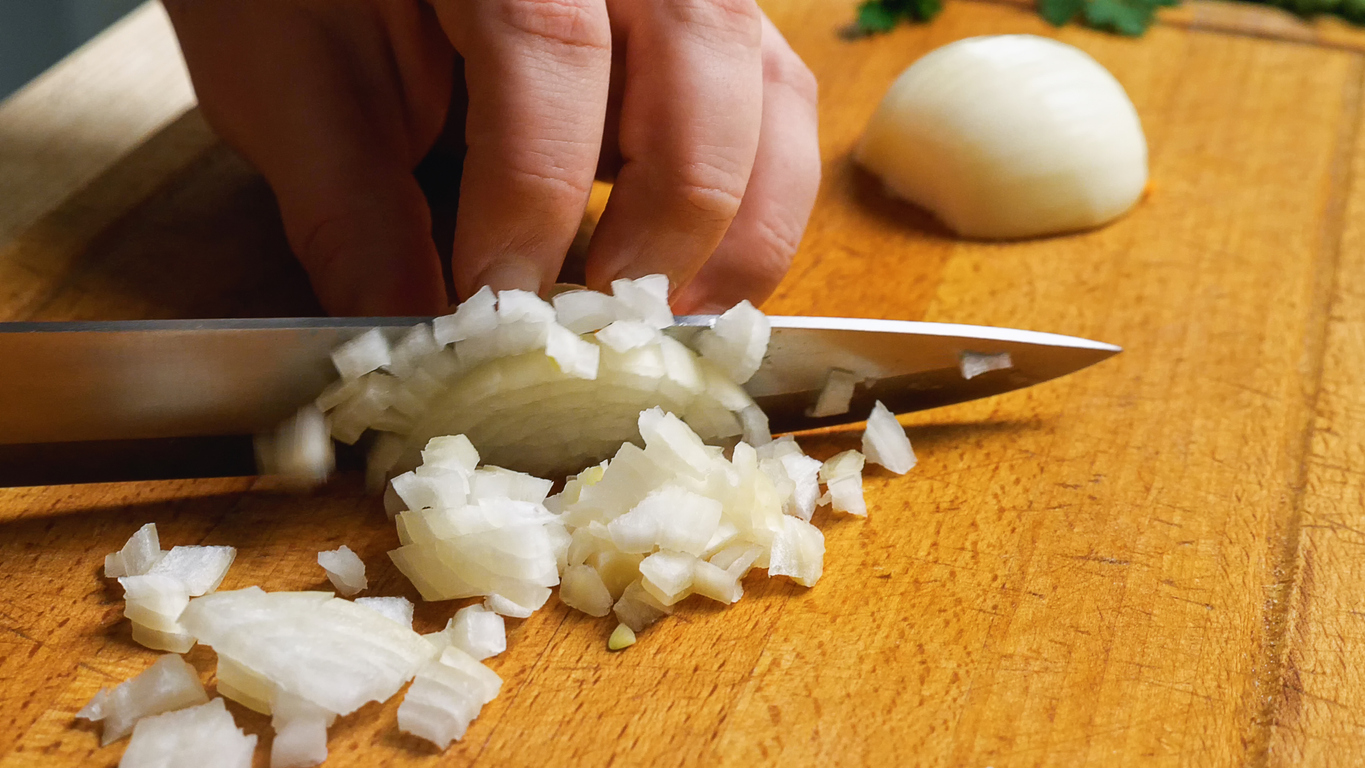 Hand würfelt eine halbe Zwiebel mit einem Messer auf einem Holzbrett.