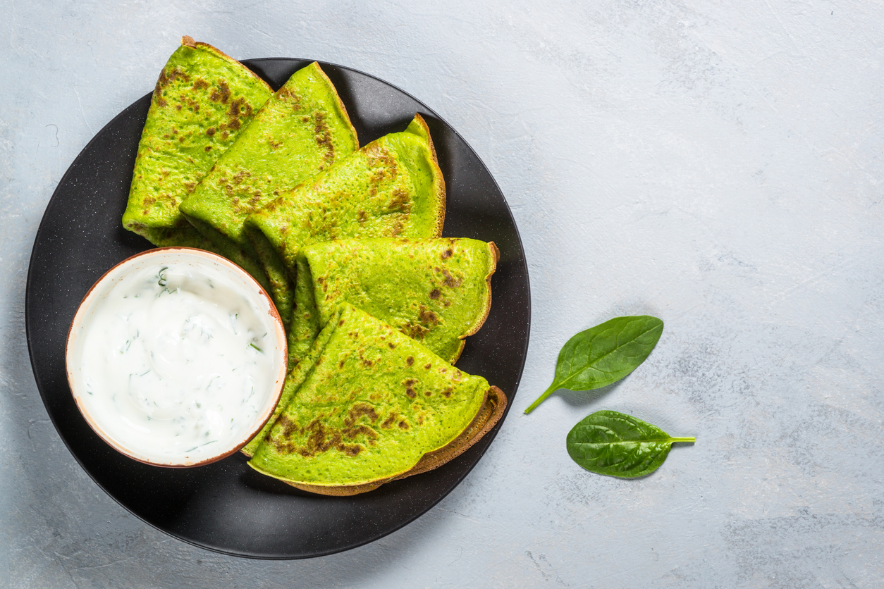 Schwarzer Teller mit mehreren gefalteten grünen Pfannkuchen und einer Schale Joghurt-Kräuterdip.