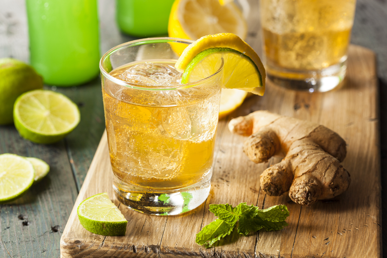 Tumbler mit Ginger Ale, Eiswürfeln und Zitronen-Limettenkeil neben Ingwer und Minze.