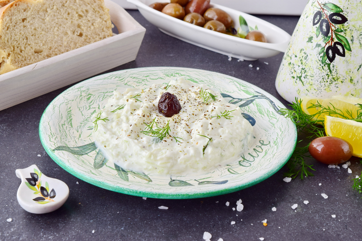 Nahaufnahme von Tzatziki mit Dill und Olive, daneben Zitronenspalten, Oliven und Brot.