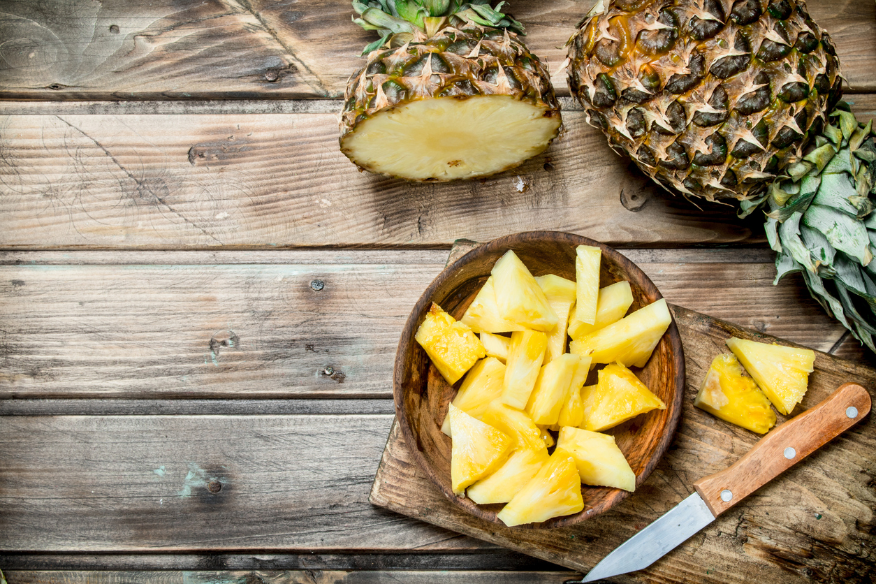 Auf einem Holztisch stehen eine Schüssel mit Ananasstücken und mehrere ganze Ananas mit Messer.