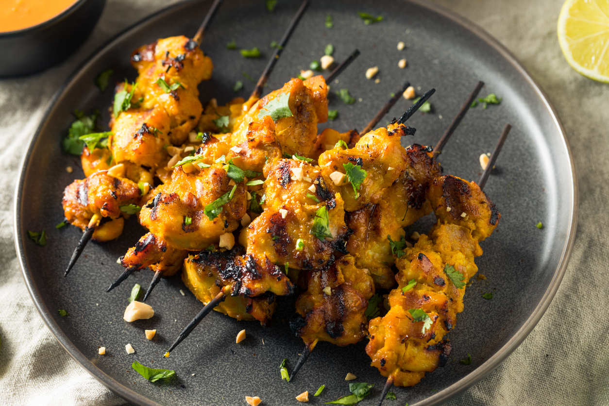 Gegrillte Chicken Satay Spieße dicht aneinander auf einem dunklen Teller mit Kräutern und Erdnussstückchen dekoriert.