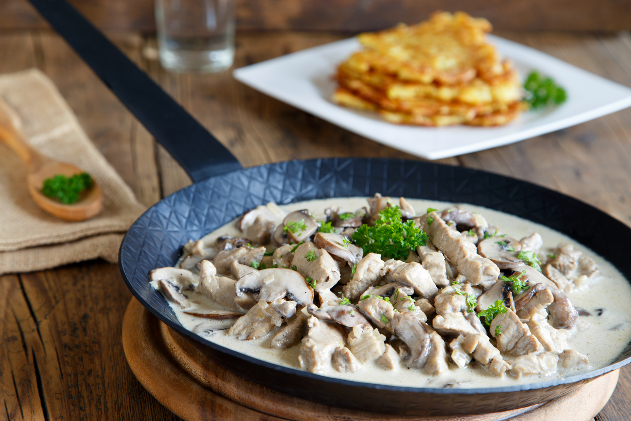 Züricher Kalbsragout mit Champignons liegt in einer Pfanne in heller Rahmsoße auf einem Holztisch.
