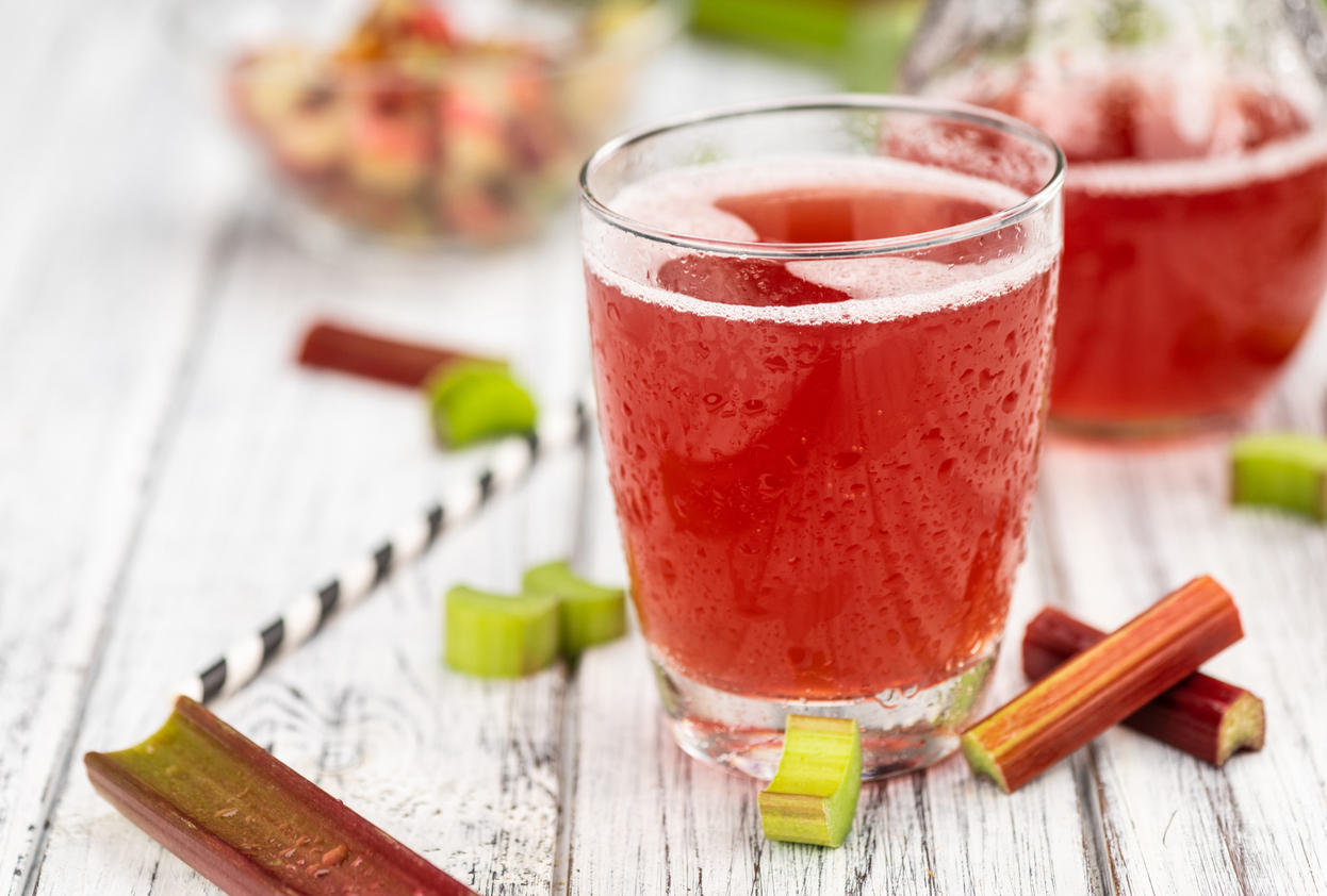 Glas mit rosa Rhabarber Limonade auf Holztisch, dekoriert mit Rhabarberstücken und gestreiftem Strohhalm.