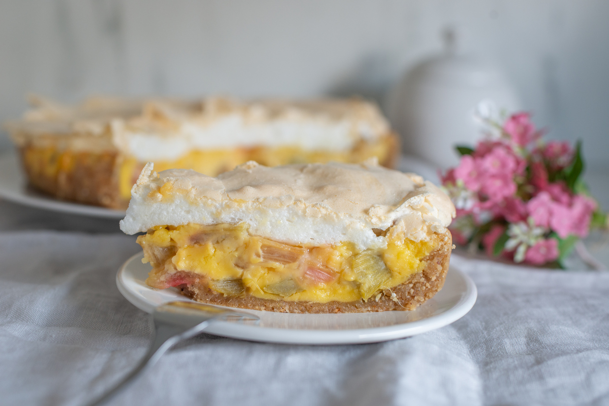 Ein Stück Kuchen mit Baiserhaube liegt auf einem Teller, dahinter die restliche Torte und rosa Blüten.