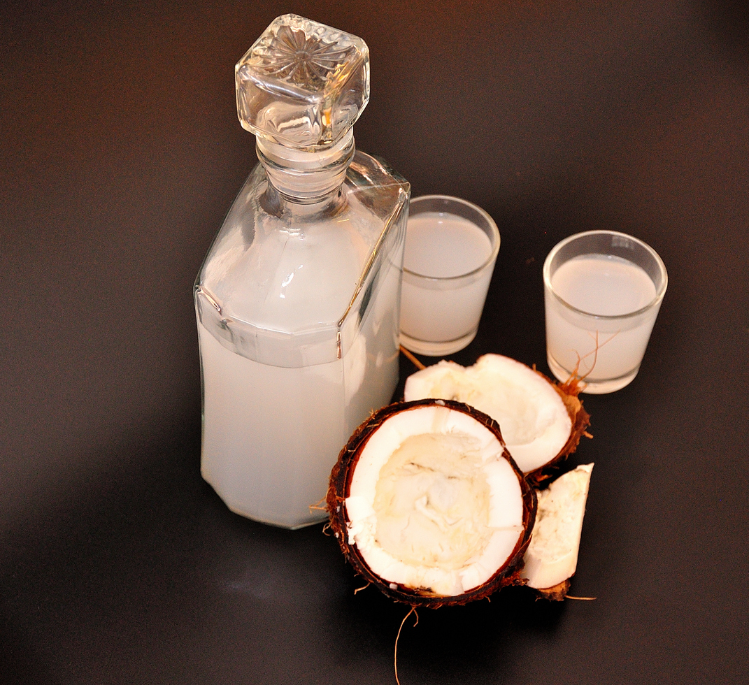 Eine Glasflasche mit Kokoslikör steht neben zwei Gläsern, davor liegen halbierte Kokosnüsse auf schwarzem Untergrund.