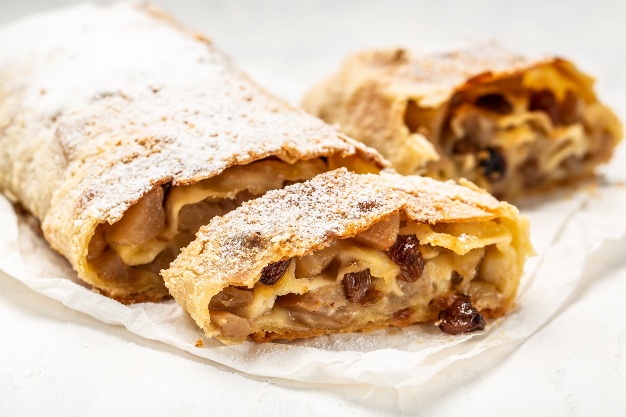 Zwei angeschnittene Stücke hausgemachter Apfelstrudel mit viel Apfel-Rosinen-Füllung liegen im Vordergrund, der restliche Strudel unscharf dahinter.