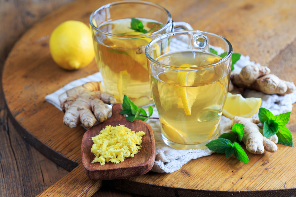Zwei Gläser Ingwertee mit Zitrone und frischer Minze stehen auf einem Holztisch, daneben liegen Ingwerwurzeln, ein ganzer Zitronen und geriebener Ingwer auf einem Holzbrett.