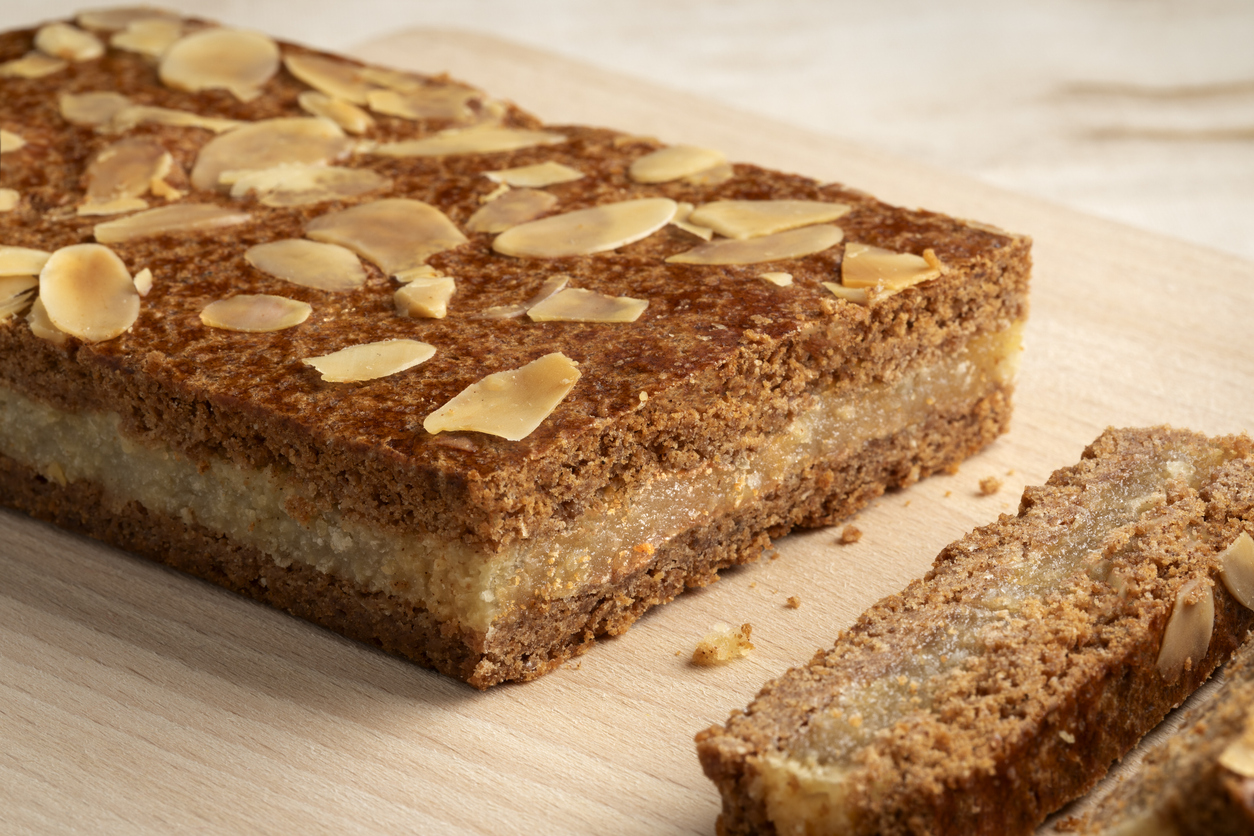 Ein länglicher Gevulde Spekulatius mit heller Füllung und Mandelblättchen liegt auf einem Holzbrett, ein Stück ist bereits angeschnitten.