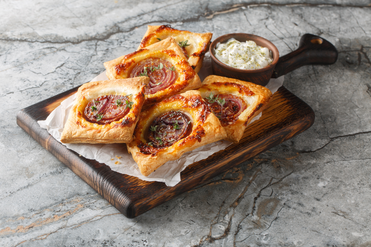 Ein Holzbrett ist mit vier Blätterteigtaschen mit karamellisierter Zwiebel und einem kleinen Schälchen Dip angerichtet.