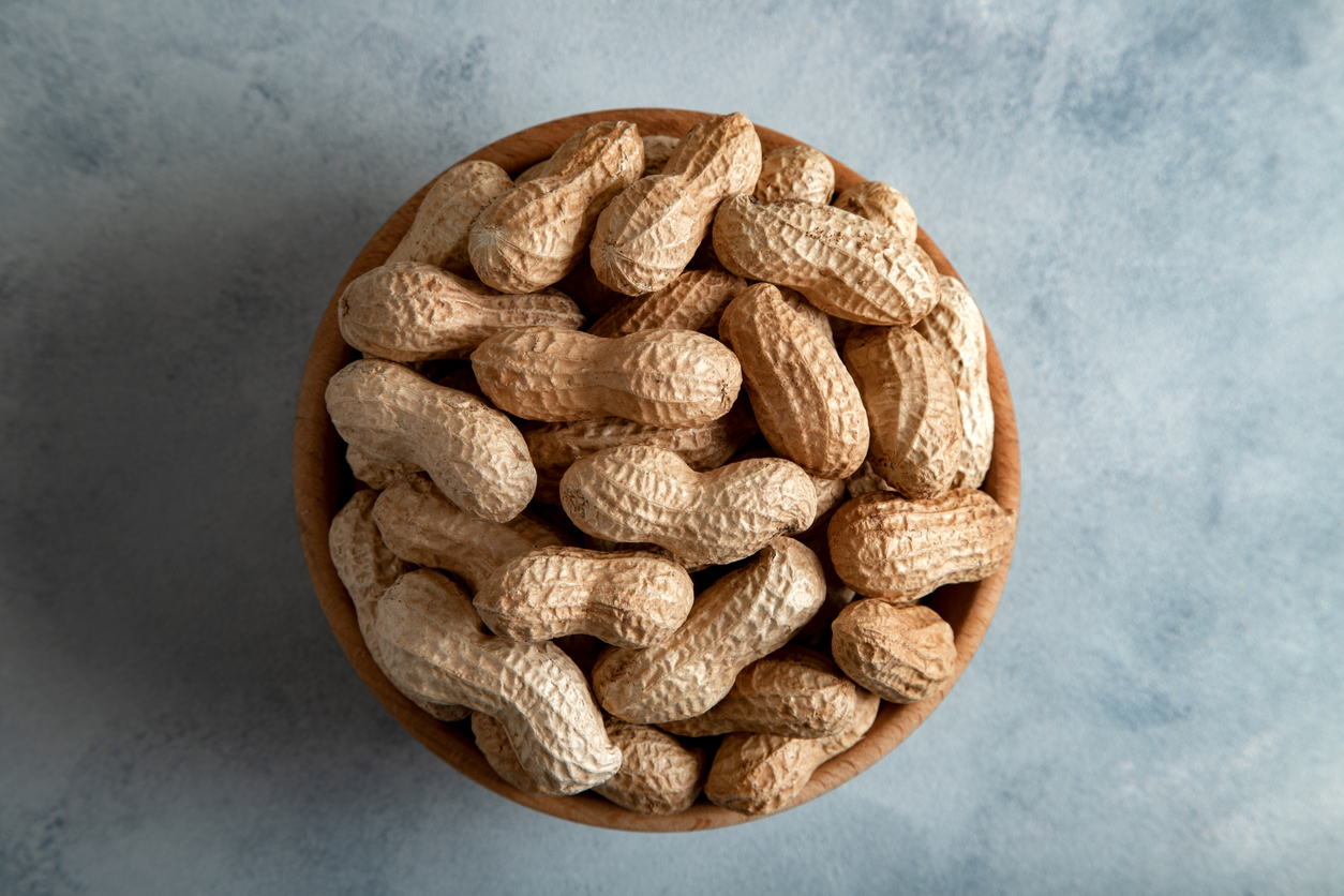 Holzschale von oben fotografiert, gefüllt mit ungeschälten Erdnüssen auf hellblauem Untergrund.
