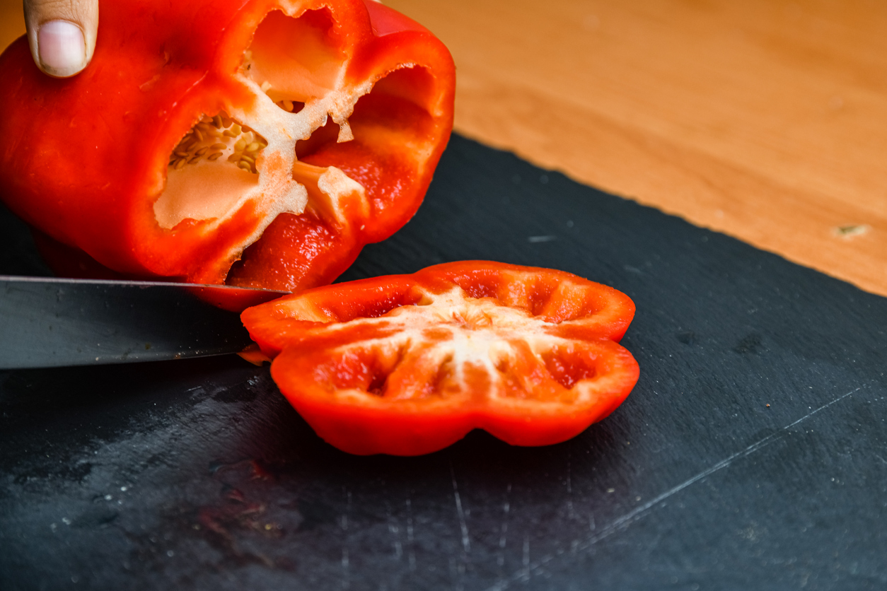 Eine rote Paprika wird auf einem dunklen Brett mit einem Messer in Scheiben geschnitten.