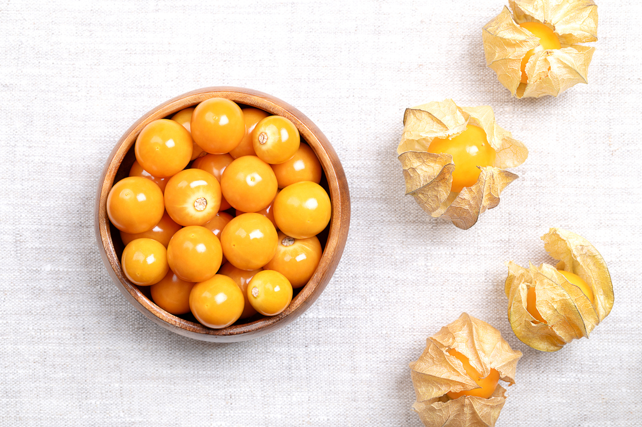 Holzschale mit vielen orangefarbenen Beeren neben geöffneten Hüllen auf hellem Stoff.
