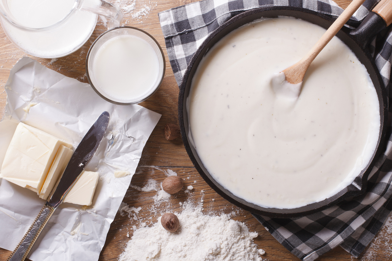 Pfanne mit frisch gekochter Béchamelsauce, daneben Milch, Butter und Mehl auf einem Holztisch.