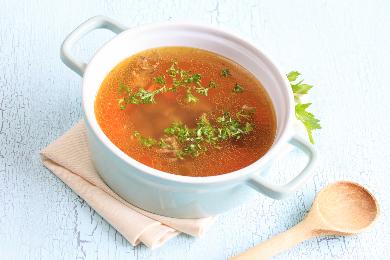 Heller Suppentopf mit klarer goldbrauner Rinderbrühe und gehackter Petersilie auf hellblauem Untergrund.