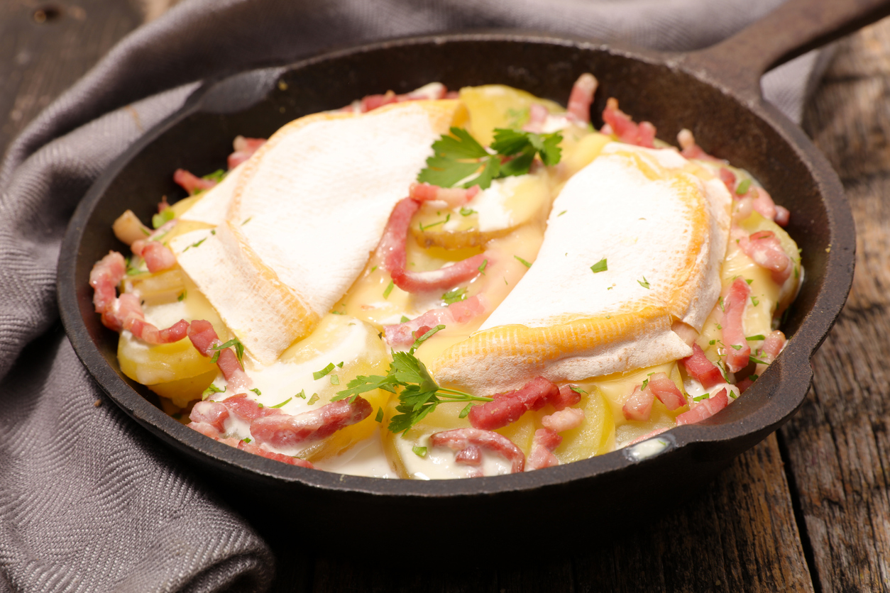 Tartiflette wird in einer schwarzen Pfanne mit Kartoffelscheiben, Speckstreifen und Käse überbacken.