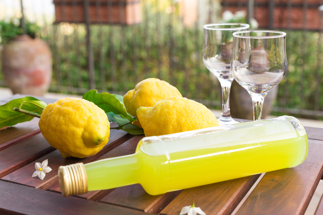 Flasche mit gelbem Limoncello liegt neben frischen Zitronen und zwei leeren Gläsern auf einem Holztisch im Freien.