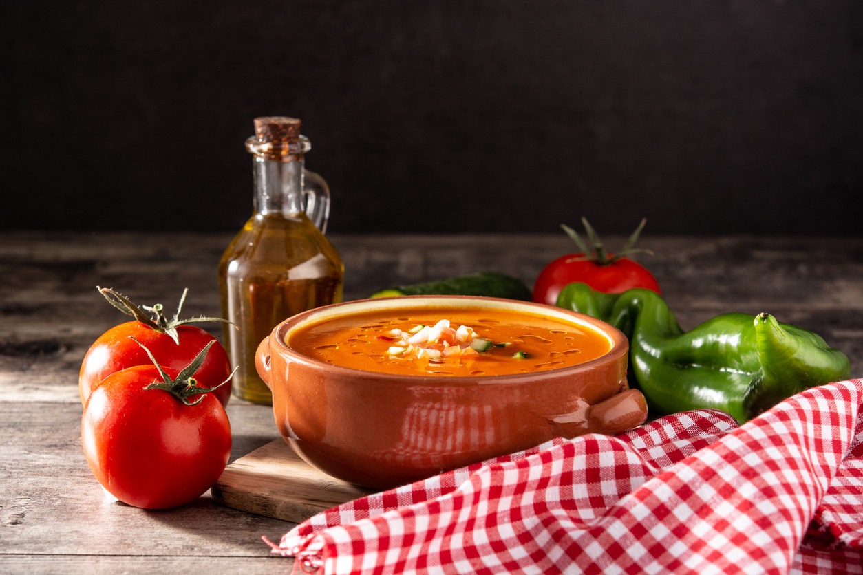 Rustikale Szene mit einer Schüssel Gazpacho, Tomaten, Paprika, Gurke und einer Flasche Olivenöl auf einem Holztisch.
