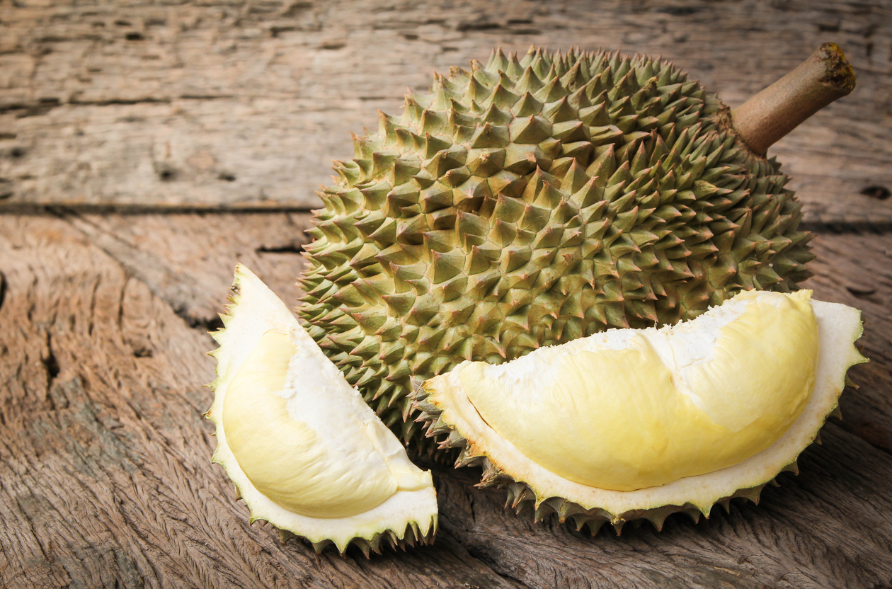 Eine ganze Durian liegt auf Holz, daneben liegt ein geöffnetes Stück mit Fruchtfleisch.