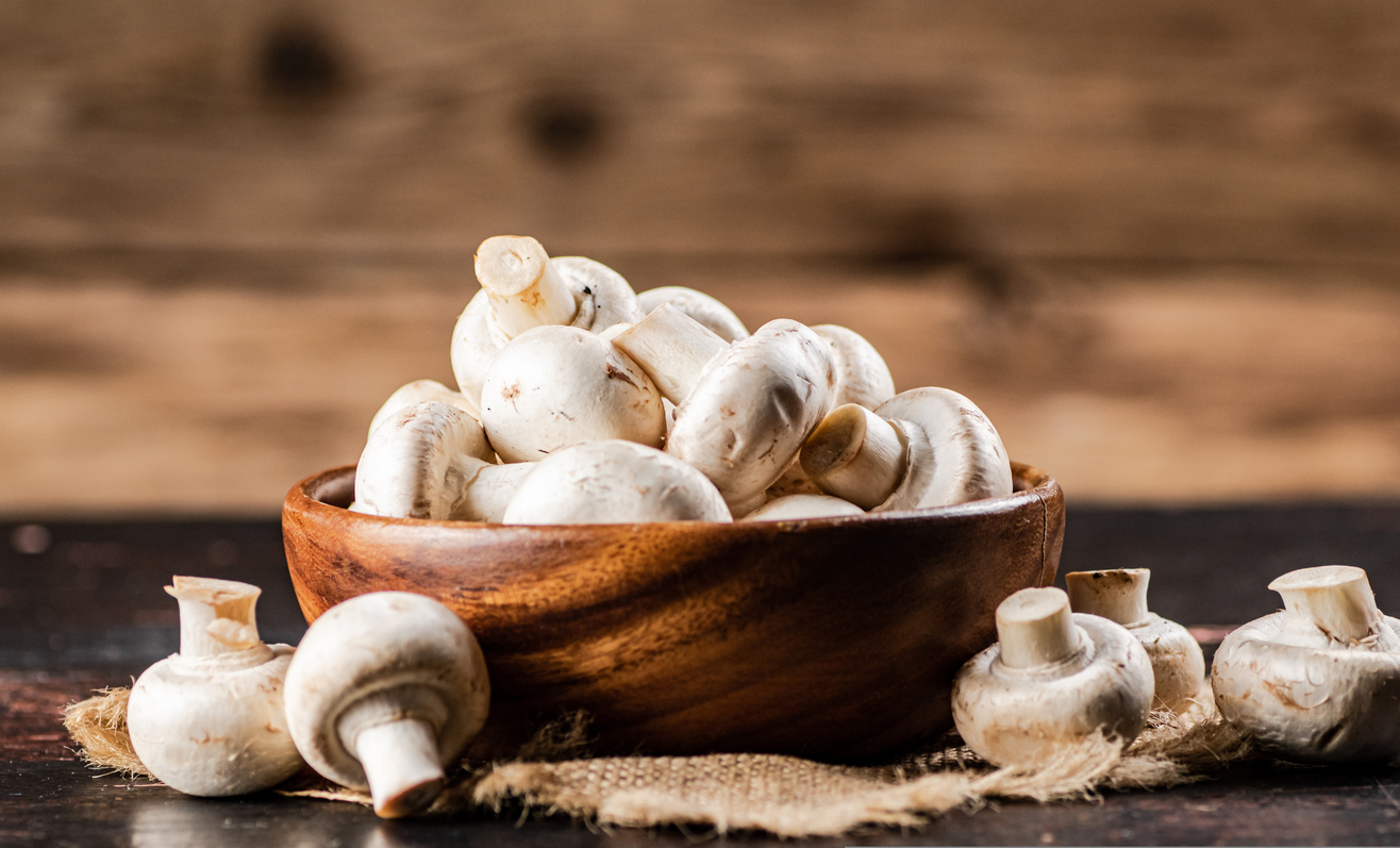 Holzschale voller weißer Champignons auf Jute vor einem Holz-Hintergrund.