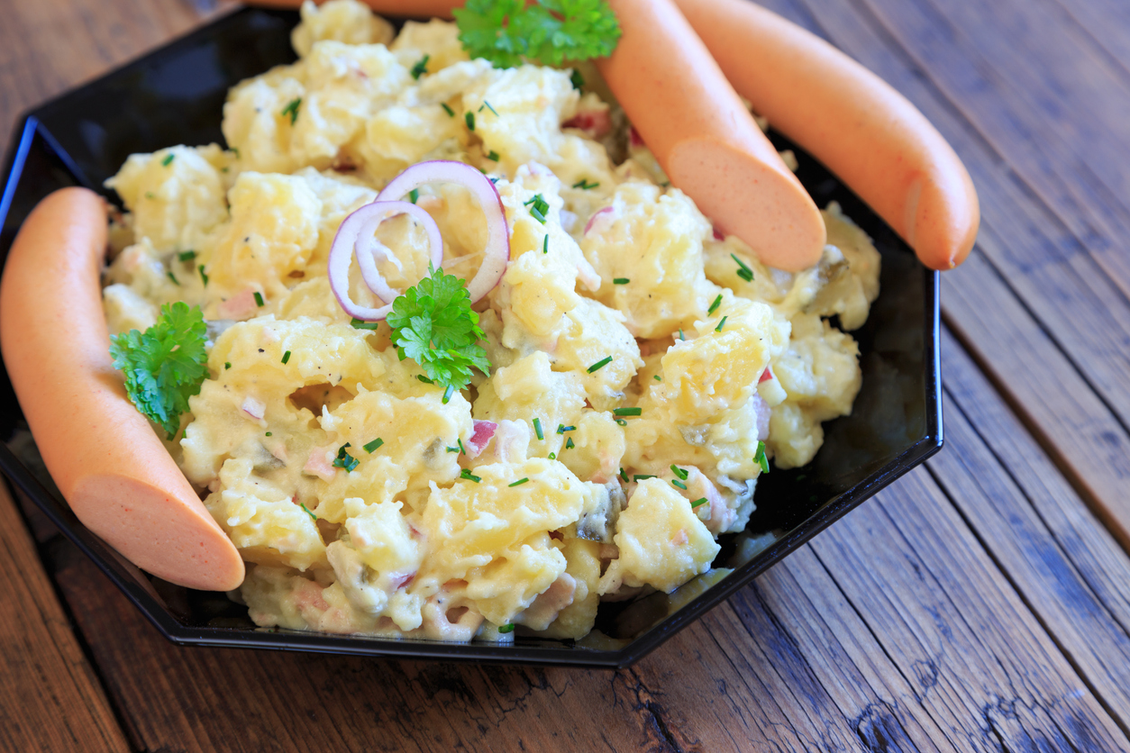 Kartoffelsalat mit Mayonnaise liegt auf einem schwarzen Teller und ist mit Kräutern und Zwiebelringen dekoriert.