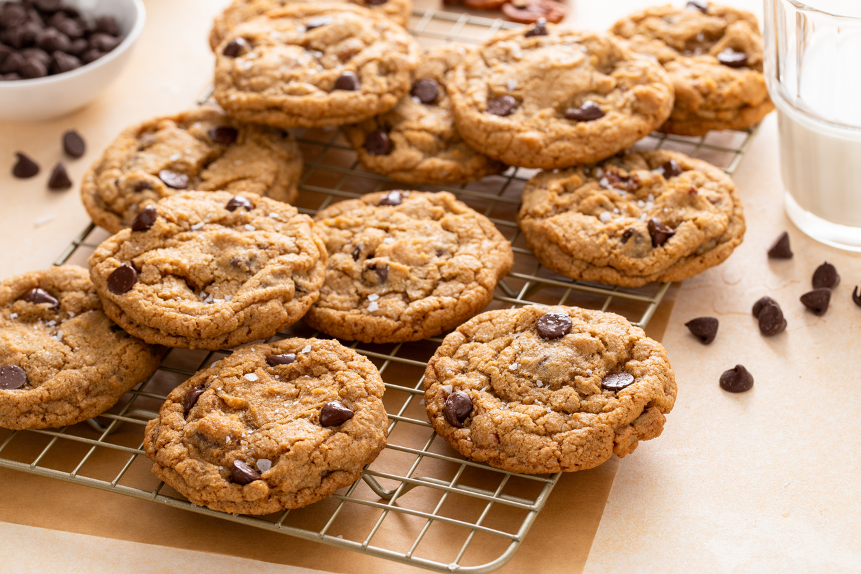 Schoko-Cookies liegen auf einem Gitter, im Hintergrund stehen Schokotropfen und ein Glas Milch.