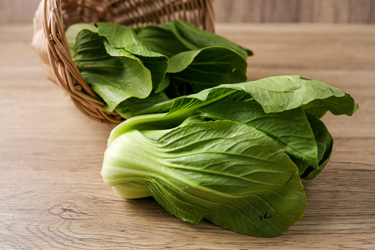 Ein Pak-Choi liegt im Vordergrund auf Holz, im Hintergrund steht ein Korb mit weiterem Pak-Choi.