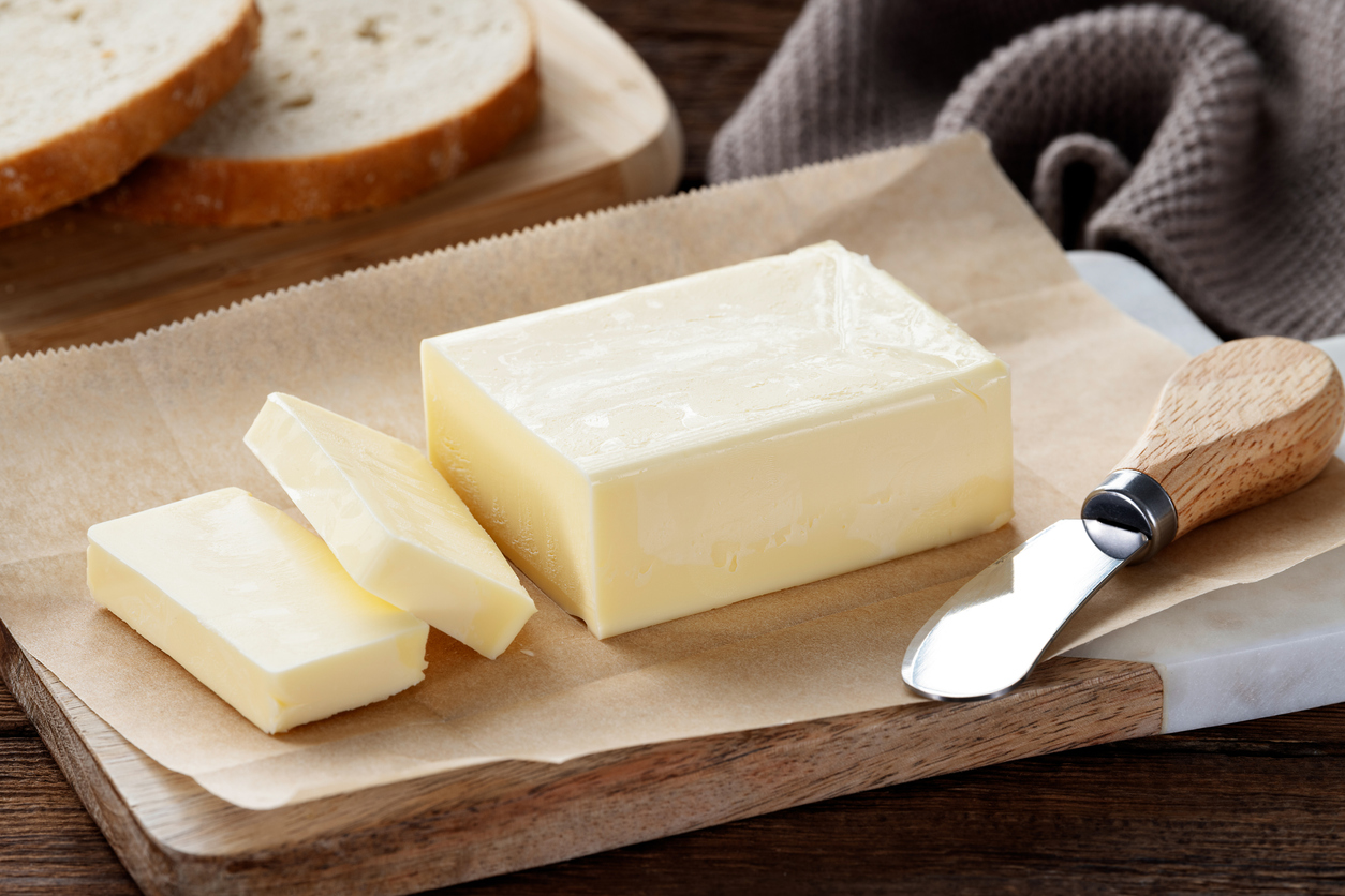 Ein Block Butter liegt auf Backpapier mit einem Buttermesser, im Hintergrund sind Brotscheiben zu sehen.