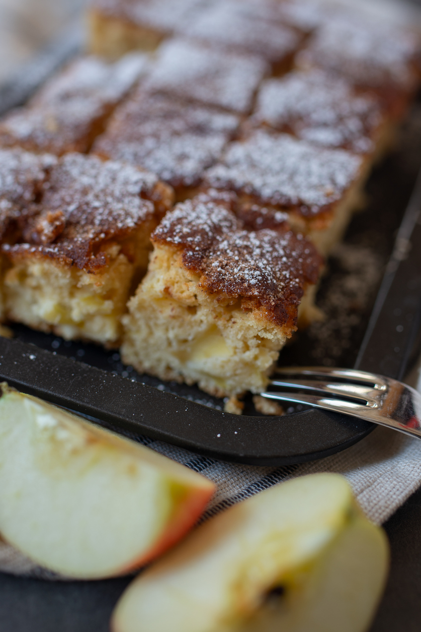 Ein Apfel-Zimt Blechkuchen ist in Quadrate geschnitten und mit Puderzucker bestäubt.