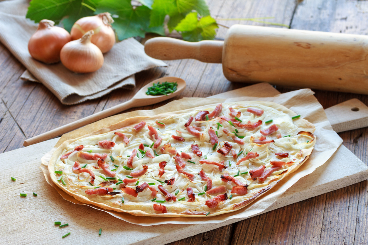 Ein Flammkuchen mit Speck und Zwiebeln liegt auf Backpapier auf einem Holzbrett neben einem Nudelholz.