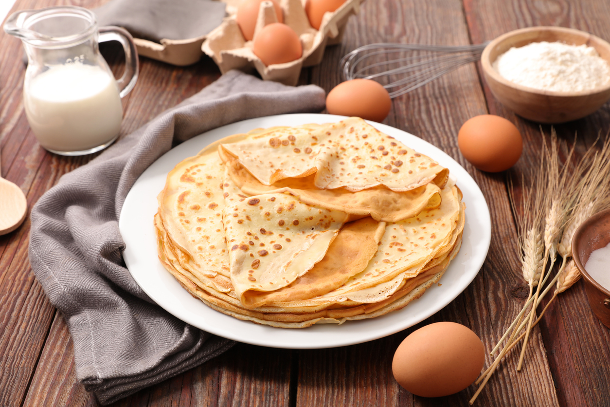 Ein Teller mit Crêpes steht auf einem Holztisch und daneben liegen Eier Milch und Mehl bereit.