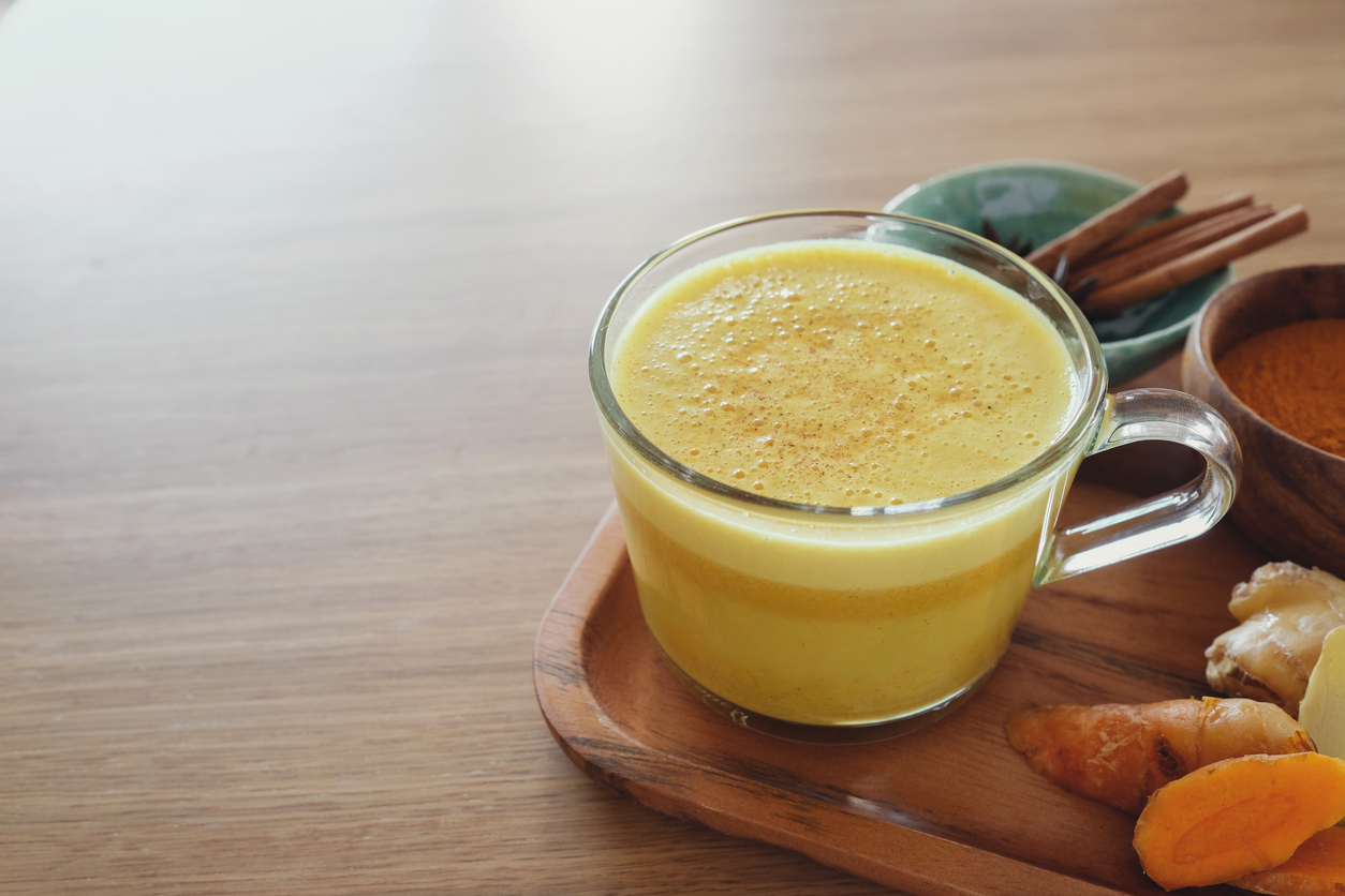 Ein Glas mit goldener Milch steht auf einem Holzbrett neben Ingwer, Kurkuma und Zimt.