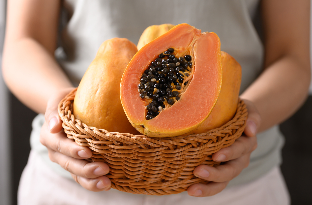 Eine Person hält einen Korb mit ganzen Papayas und einer aufgeschnittenen Papaya mit schwarzen Kernen.