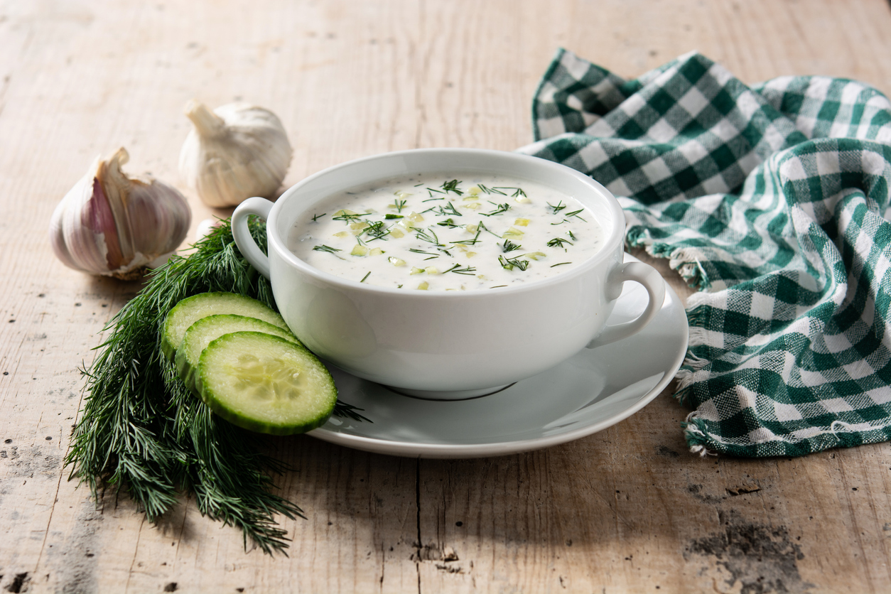 Eine weiße Tasse Tarator Sauermilchsuppe steht auf einer Untertasse neben Gurke, Dill und einem karierten Tuch.