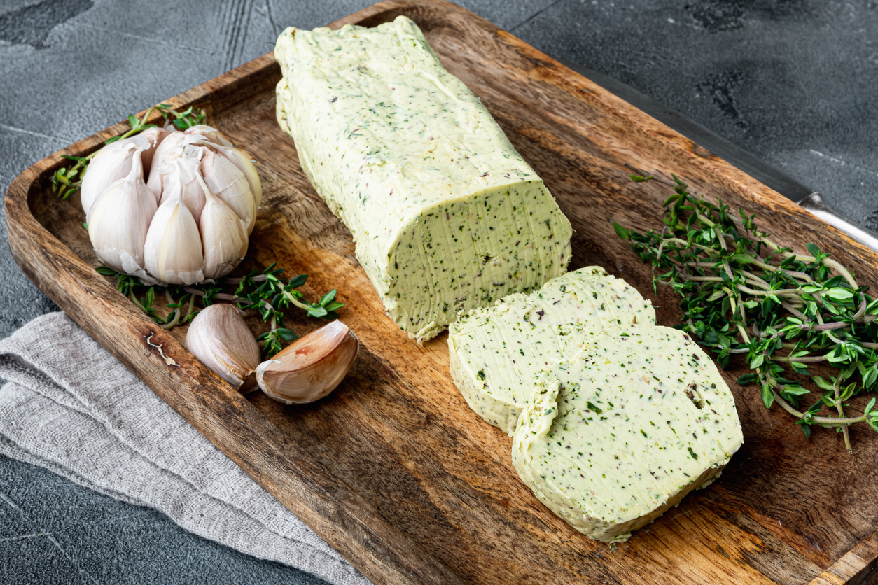 Eine Rolle Kräuter Knoblauchbutter liegt auf einem Holzbrett neben Knoblauchzehen und Thymian.