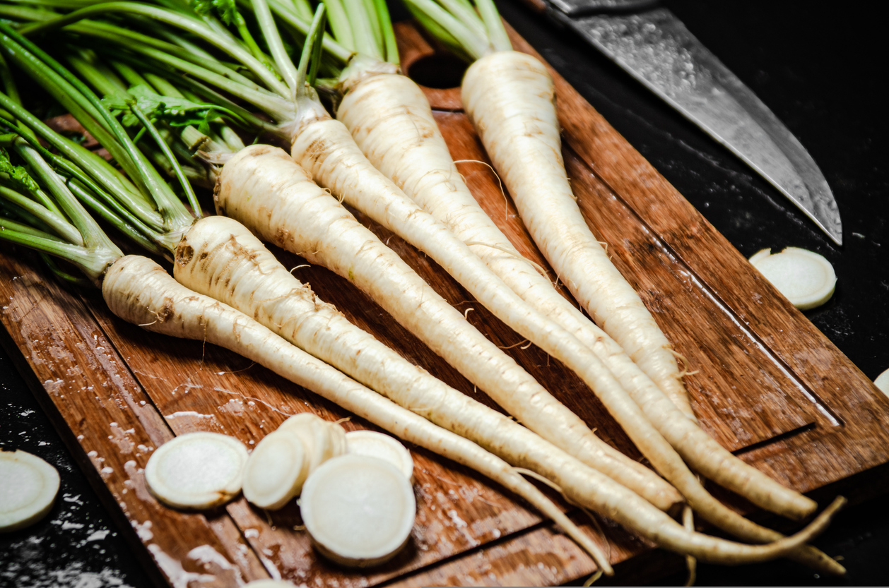 Petersilienwurzeln liegen auf einem Holzbrett neben einem Messer, dazu sind geschnittene Scheiben zu sehen.