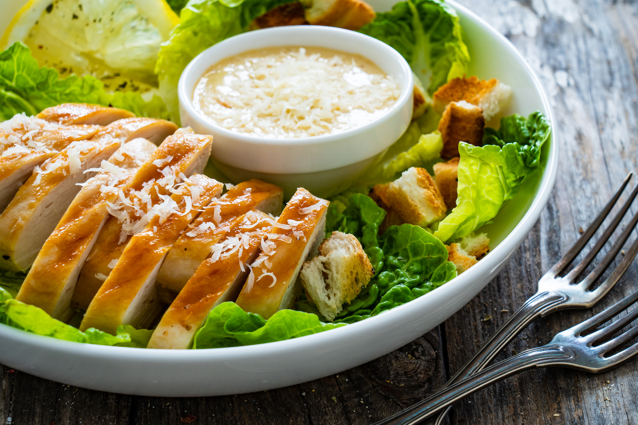 Ein Caesar Salat mit Hähnchen, Croutons und Parmesan liegt in einer Schüssel neben Besteck auf einem Holztisch.