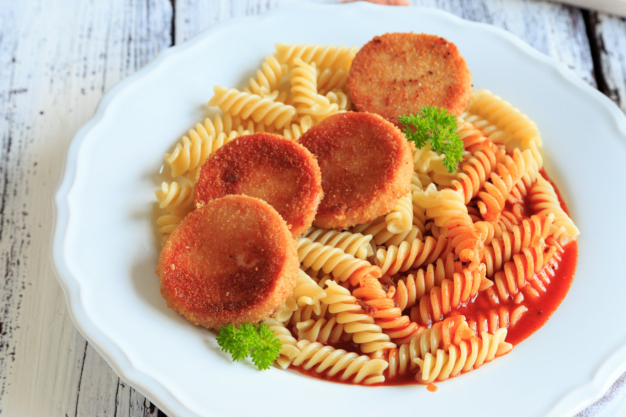 Drei panierte Jagdwurstscheiben liegen auf Spirelli mit Tomatensoße und Petersilie auf einem weißen Teller.