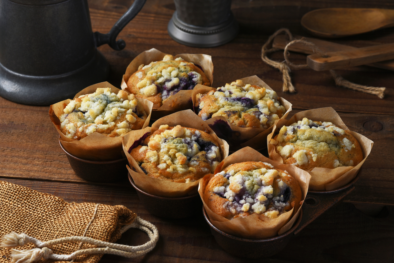 Mehrere Blaubeermuffins mit Streuseln stehen in Förmchen auf einem dunklen Holztisch.
