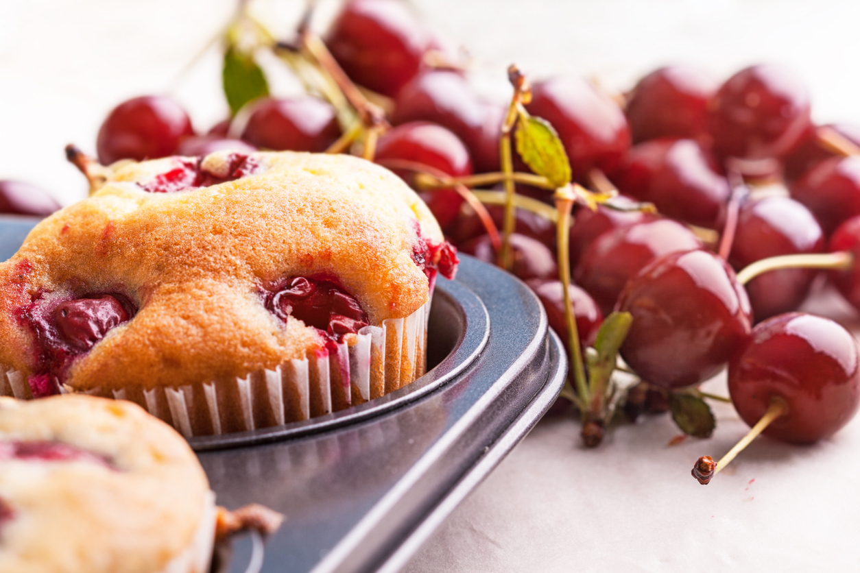 Ein Kirschmuffin sitzt in einer Muffinform, im Hintergrund liegen frische Kirschen.