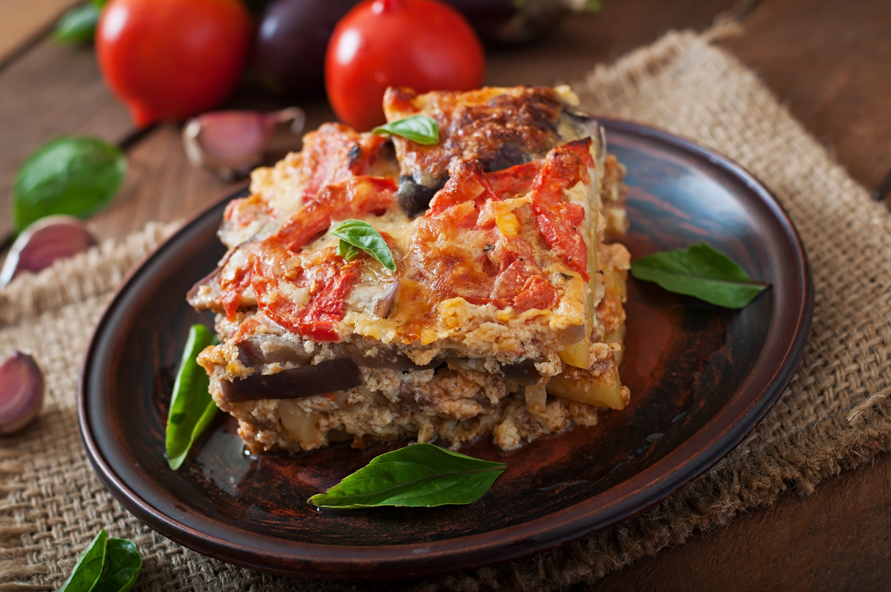 Ein quadratisches Stück Moussaka steht auf einem Teller, im Hintergrund liegen Tomaten und Auberginen.
