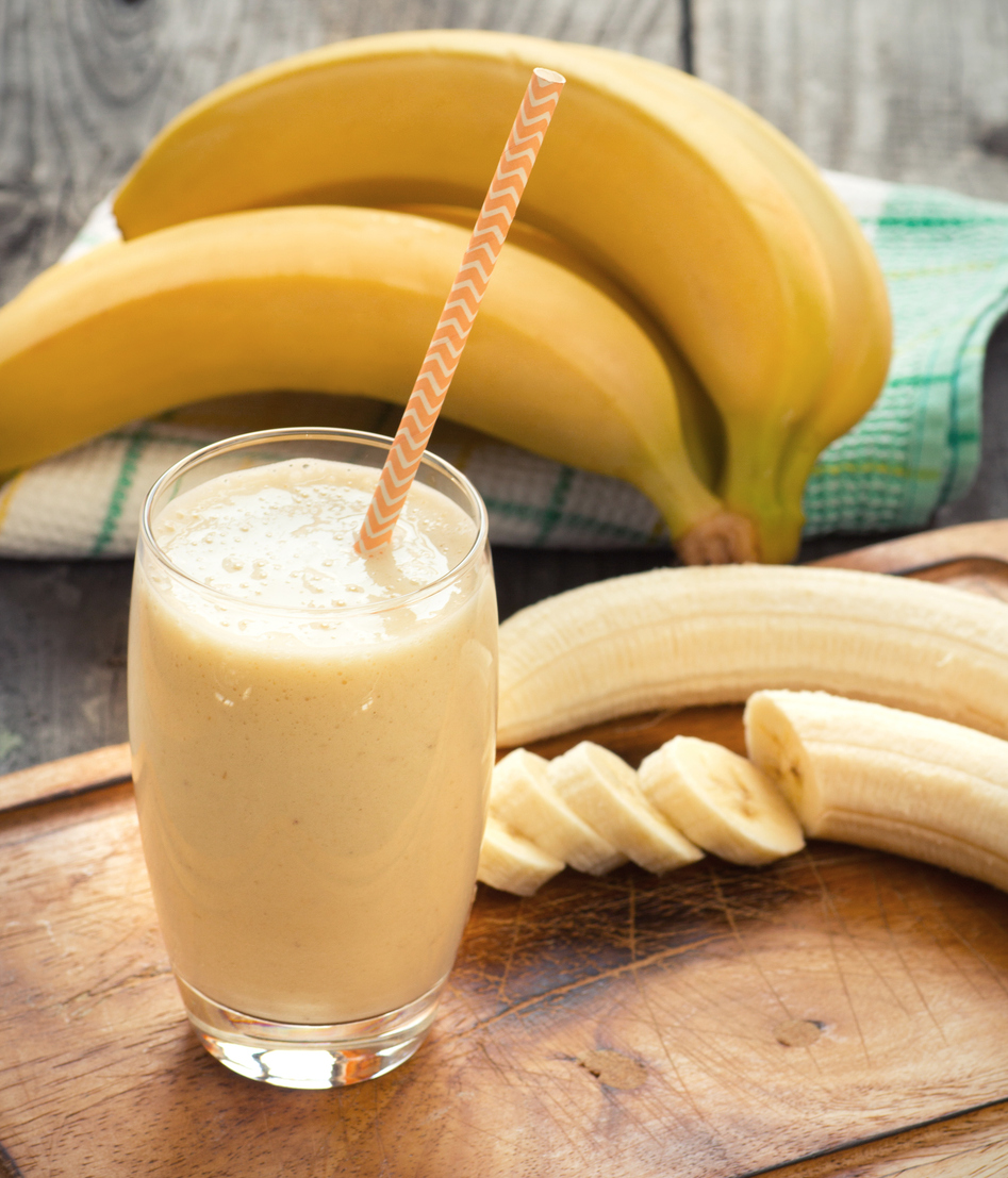 Ein Bananenshake im hohen Glas mit gestreiftem Strohhalm steht vor mehreren Bananen.