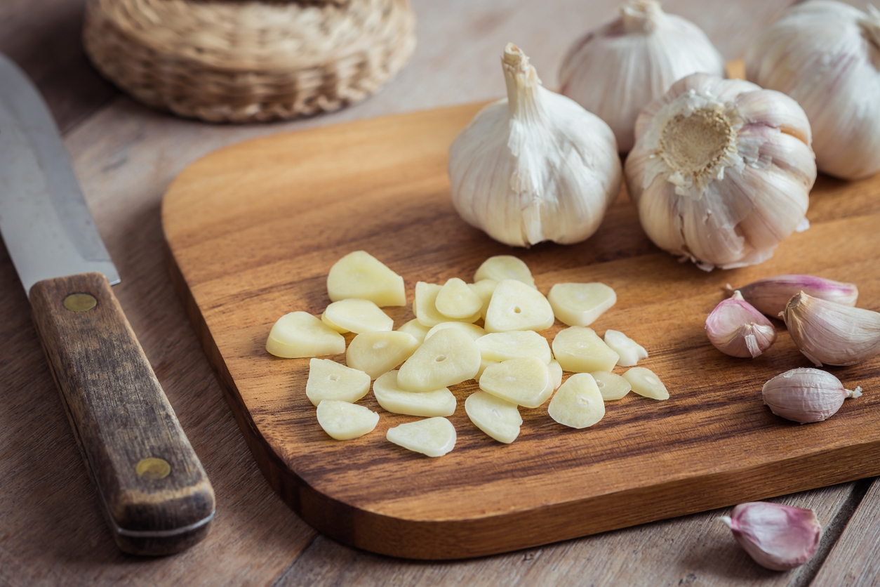 Geschnittener Knoblauch liegt auf einem Holzbrett, daneben liegen ganze Knollen und ein Messer.
