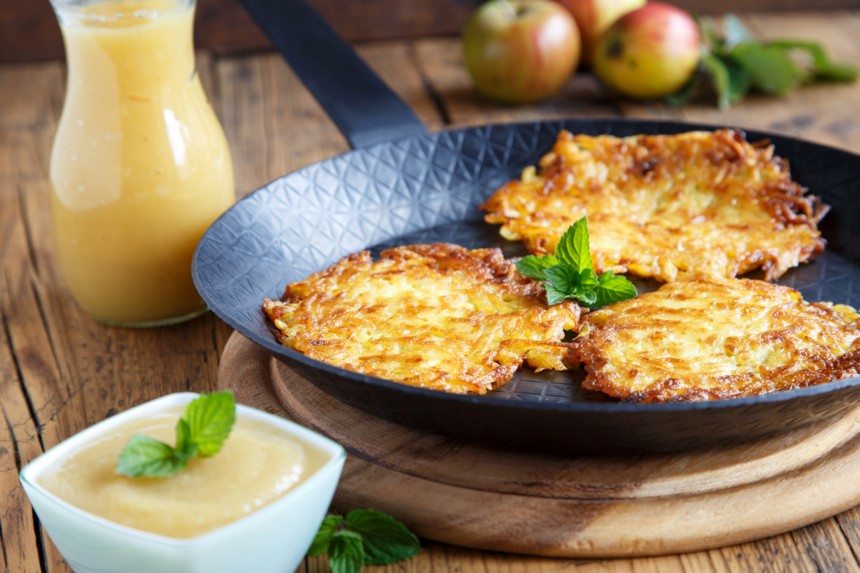 Mehrere Kartoffelpuffer liegen in einer Pfanne, daneben stehen Apfelmus und ein Krug Apfelsoße auf dem Tisch.