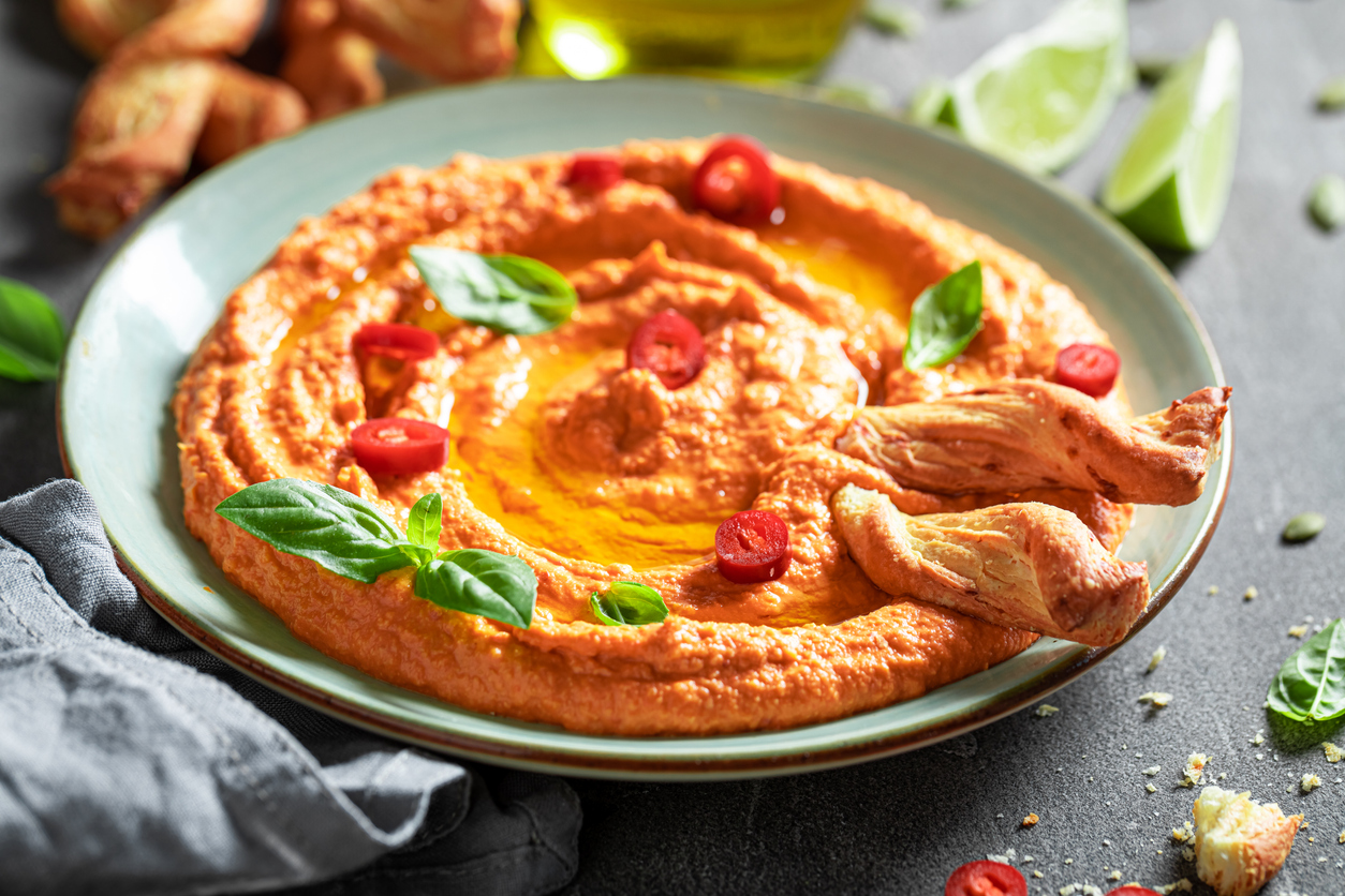 Ein hellblauer Teller mit orangefarbenem Hummus, garniert mit frischen Basilikumblättern, roten Chiliringen und knusprigen Gebäckstangen.